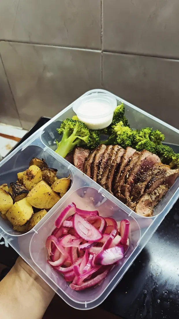 A three-compartment meal prep box with sliced beef, broccoli, potatoes and pickled onion