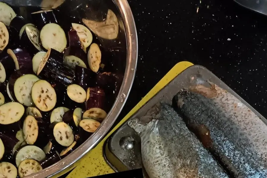 Sliced aubergine in a pot beside seasoned fish fillets on a board